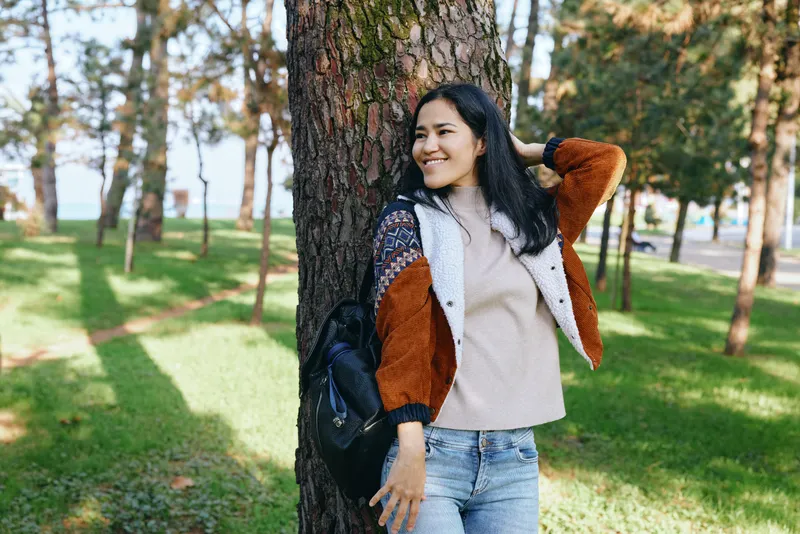Smiling woman leaning against a tree in a sunlit park, casual outfit with sweater and jacket, relaxed mood, outdoor leisure, nature scene, greenery and shadows, perfect moment for lifestyle and calm