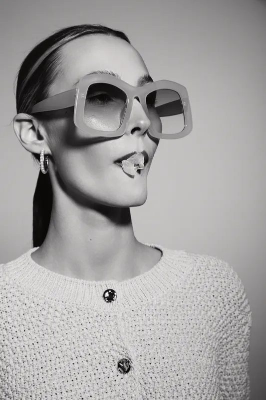 Stylish white woman with pale skin wearing oversized sunglasses and knit sweater poses with tongue out in black and white studio portrait.