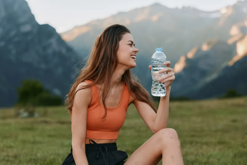 A cheerful young woman sits outdoors in a scenic field, holding a water bottle and smiling. She wears a bright sport top and shorts, enjoying a sunny moment in nature, radiating energy, health, and