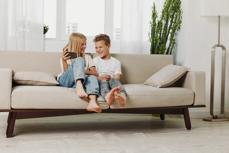 Two barefoot children sit on a beige sofa in a bright living room sharing a smartphone and laughing. Casual home scene with natural light and cozy atmosphere, concept of childhood and technology.
