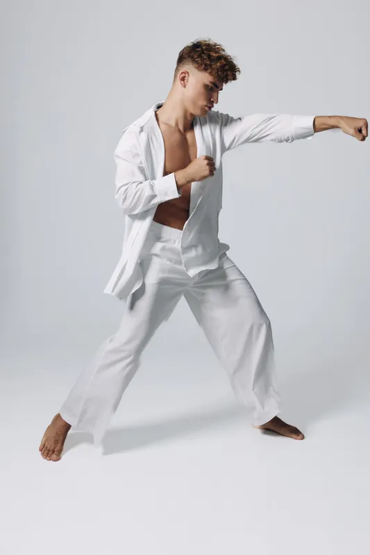Young man practicing martial arts in white loose clothing, performing a punch with focused expression on plain background. Barefoot athlete in dynamic stance for training or sport.