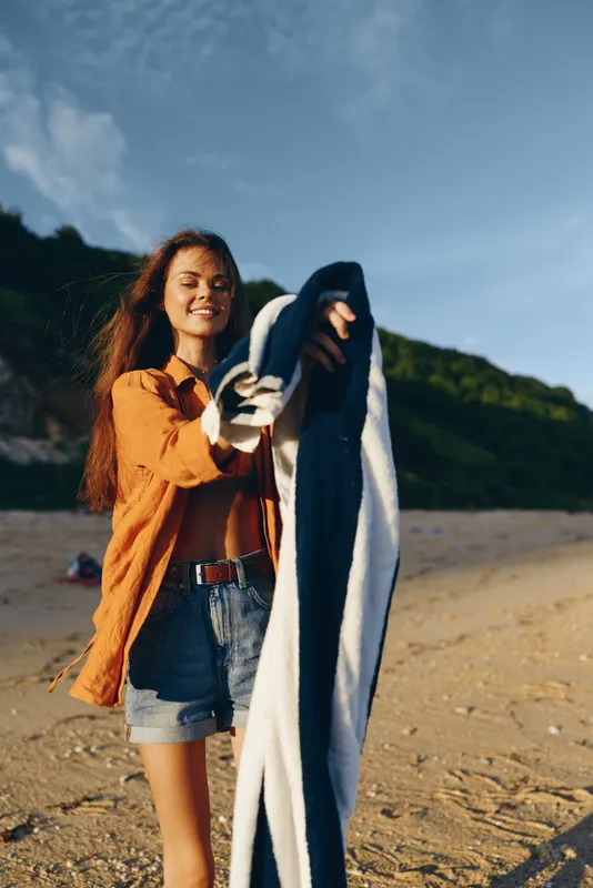 Woman smiling on beach outdoors holding jacket, warm sunlight, sandy shore and calm sea waves behind, green hills in distance, relaxed casual summer travel mood