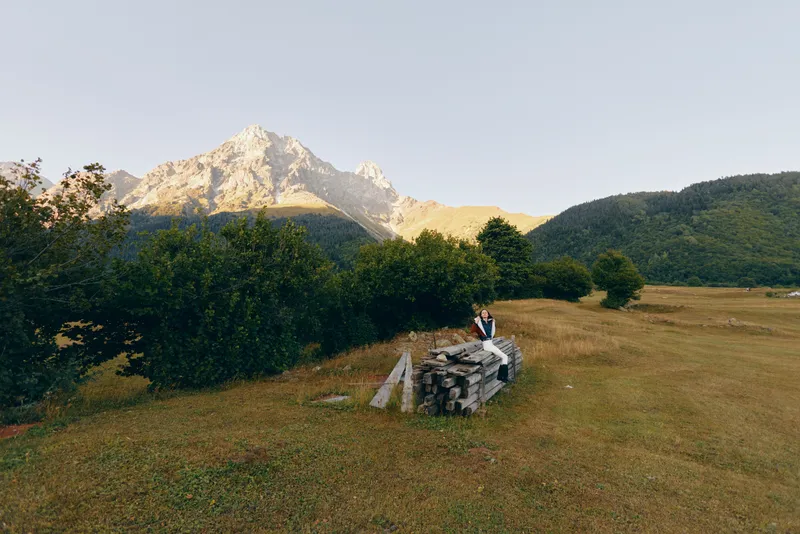 Mountain meadow with hiker and backpack sitting on a stonewall, trees and distant peaks at sunrise. Landscape scene of nature, solitude, outdoor travel and hiking adventure.