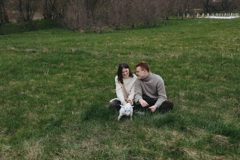 Couple with a white goat kid in a grassy field, sharing a gentle moment. Sunlit rural scene conveys lifestyle and affection, natural joy, and relaxed, candid connection between people and animals.
