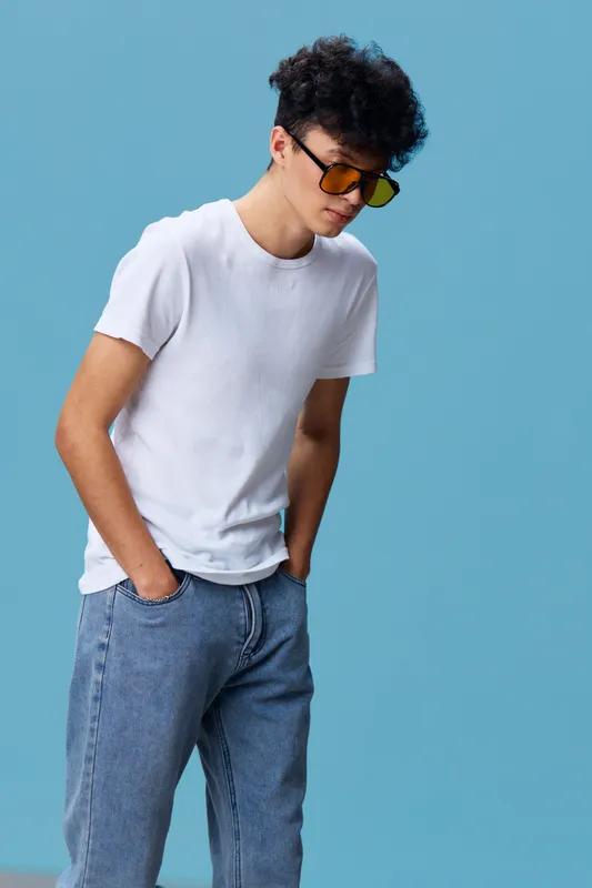 young man wearing sunglasses and white t-shirt posing casually with hands in pockets against blue background, stylish and relaxed urban look