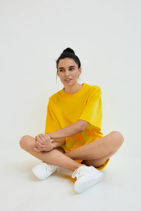 Young woman wearing bright yellow oversized t-shirt sitting cross-legged on a white floor, casual style, relaxed pose, simple studio background, warm natural lighting.
