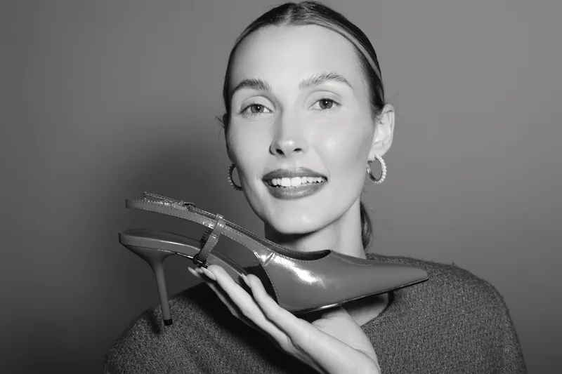 Young white woman with fair skin smiles holding high heel shoe near face, isolated on grey background. Concept of fashion, beauty, footwear, and style in studio lighting.
