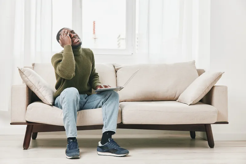 A frustrated young African American man sitting on a beige sofa, holding his head while working on a laptop, showcasing stress in a modern living room