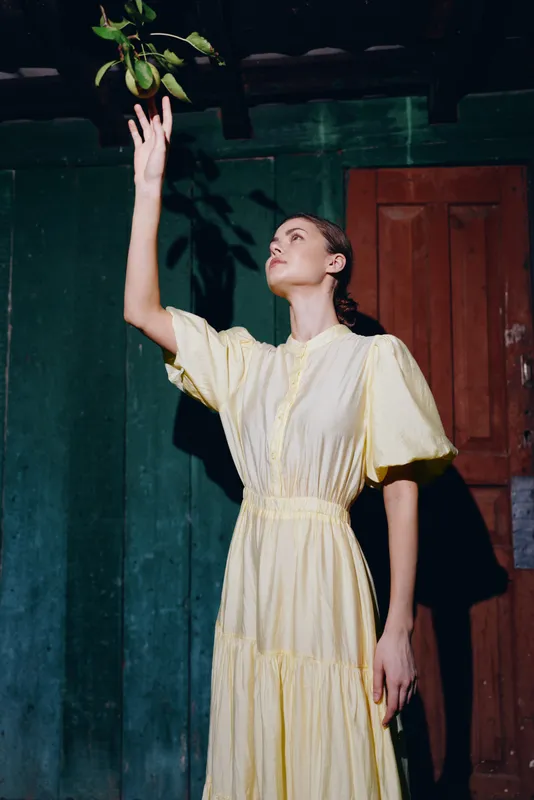 woman dress reaching fruit portrait sunlight shadow, yellow vintage dress and wooden wall, contemplative pose with natural light, authenticity inclusivity and timeless simple elegance