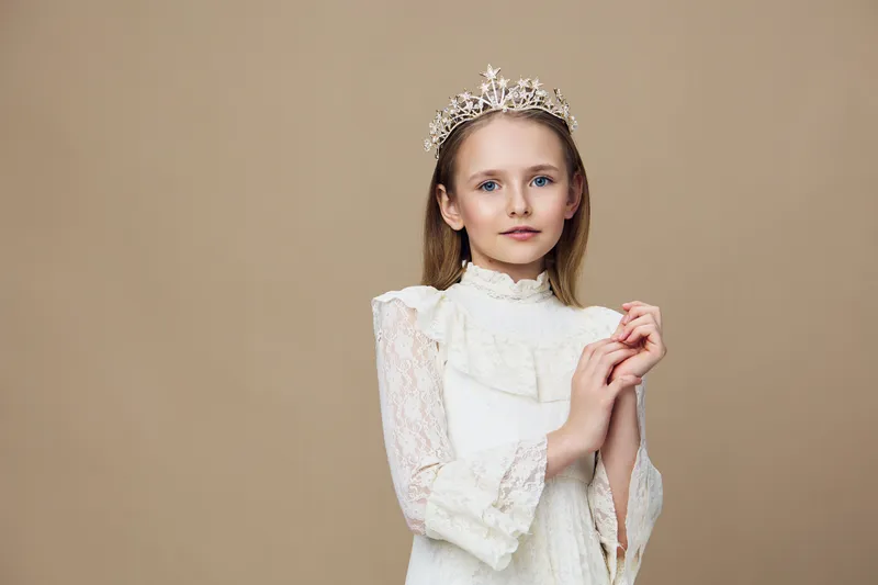 Young girl wearing a crown, dressed in a white vintage gown, with a serene expression against a neutral colored background, perfect for children s portraits