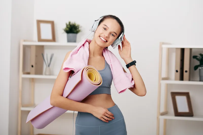 Fitness woman in athletic wear with yoga mat, headphones and pink towel, smiling during a workout in a bright home studio with shelves, minimalist decor and natural light.
