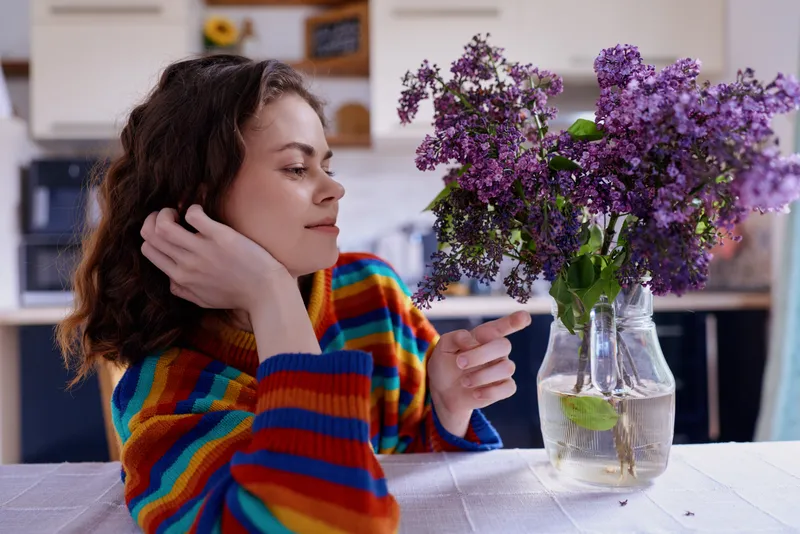 Young woman with curly hair wearing a colorful striped sweater sits at a kitchen table, gazing thoughtfully at a vase of purple flowers, creating a cozy home mood and calm atmosphere.