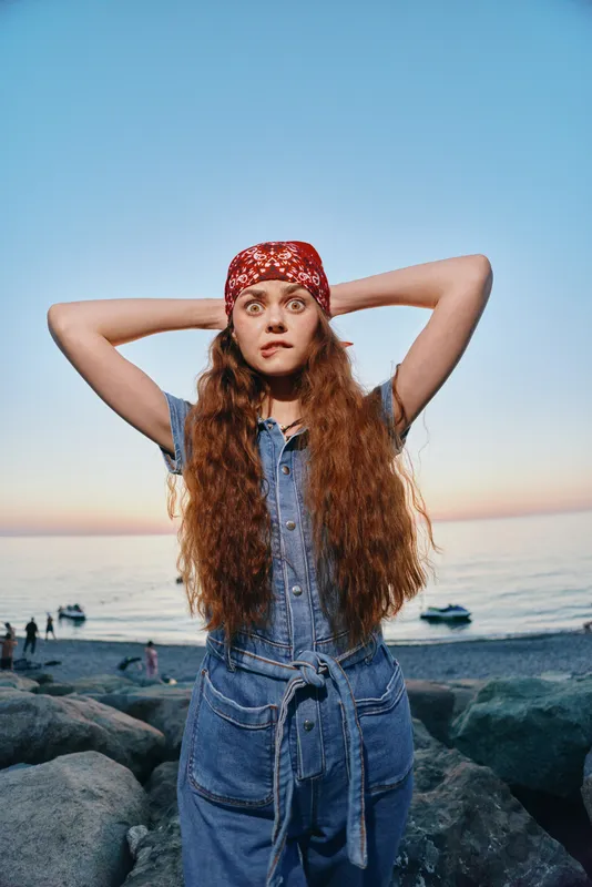 young woman with long hair wearing red bandana and denim dress standing on rocky beach at sunset with calm sea and soft sky