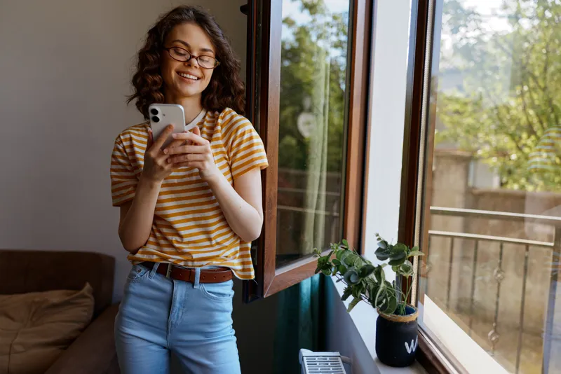 Smiling woman with striped shirt using smartphone by an open window at home, bright natural light, casual lifestyle, cozy interior, cheerful mood, everyday moment.