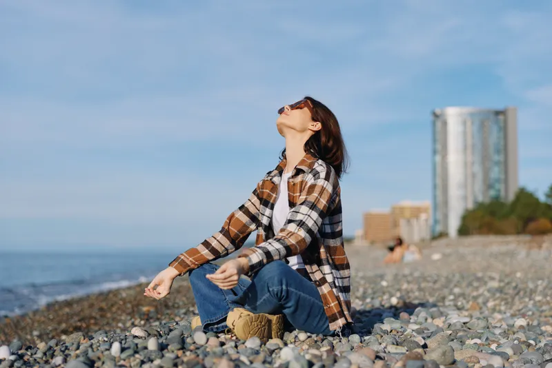 Woman sitting on a pebble beach in plaid shirt and jeans, gazing at the sky with a calm expression, sea breeze and sunlight creating a serene outdoor moment with distant skyline