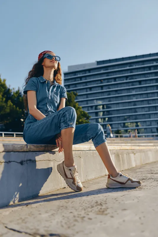 lifestyle woman in modern boho-western denim street style sitting outdoors under film color with sunglasses and casual sneakers on sunny day