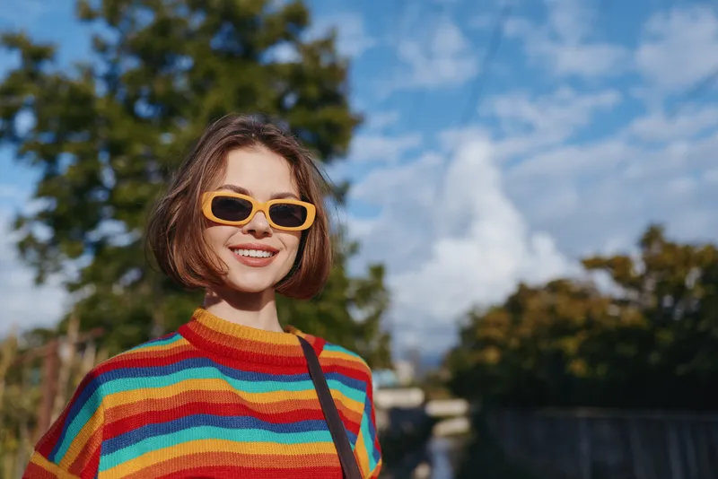Woman smiling in a colorful rainbow sweater and orange sunglasses, short bob haircut, outdoor portrait by a canal with trees and blue sky, casual shoulder bag, sunny travel lifestyle.
