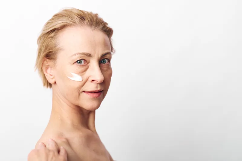Mature woman with short blonde hair applies white face cream on cheek, looking at camera. Skincare concept for aging skin, self-care, beauty routine, and healthy lifestyle on white background.