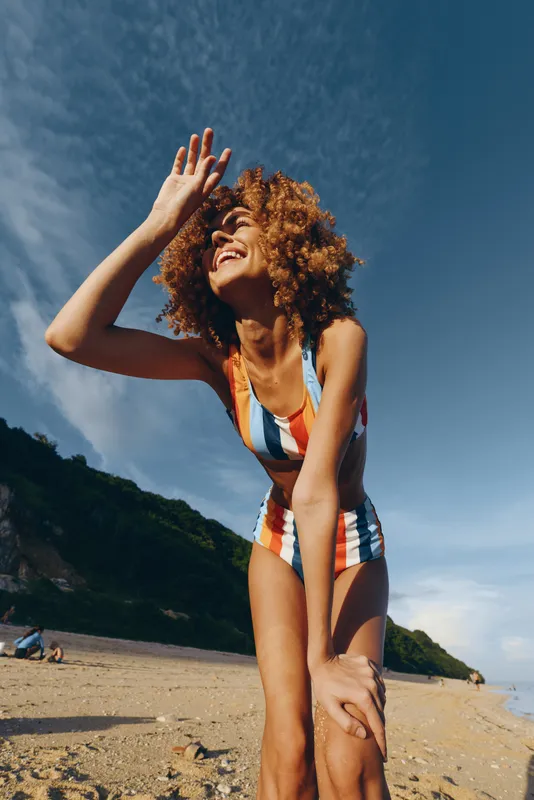 Young woman with curly hair wearing a striped swimsuit enjoys sunny weather on a sandy beach while smiling and shielding her eyes from the bright sky.