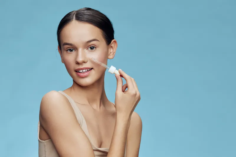 Young woman applying facial serum on a blue background, showcasing healthy skin and beauty routine with a cheerful expression, perfect for skincare concepts
