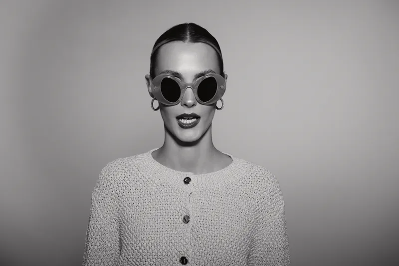 Stylish Caucasian woman with pale skin wears round sunglasses and textured sweater, posing confidently against plain background. Concept of fashion and attitude.