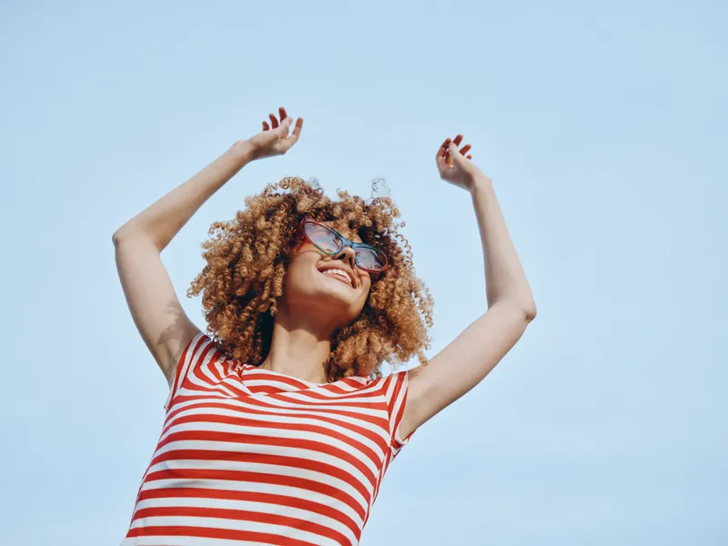 Young woman with curly hair in a red and white striped top raises her arms in joy under a clear blue sky, capturing carefree summer energy and vibrant outdoor vibes.
