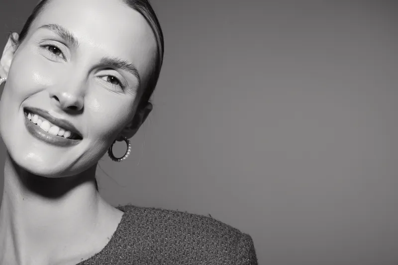 Smiling young caucasian woman with light skin and short hair poses in studio with plain background, wearing textured top and hoop earrings.