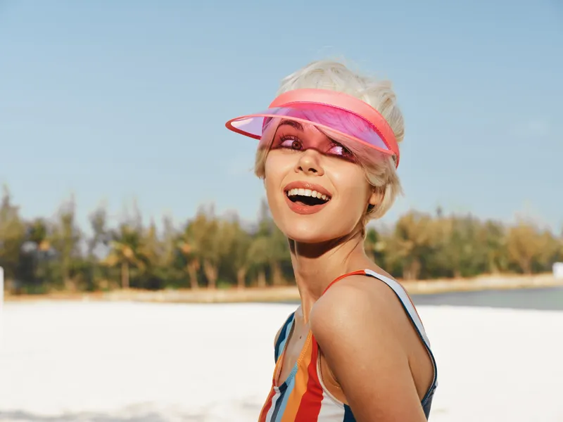 Smiling woman on a sunny beach, wearing a striped swimsuit and a pink visor, sunlit skin and a bright, carefree expression in a vivid outdoor portrait.