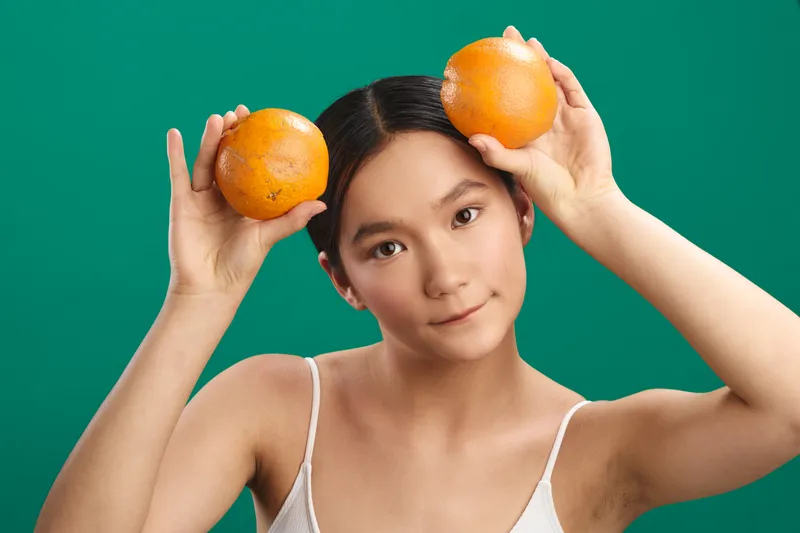 asian woman holds oranges near her head in a vibrant studio setting, smiling softly at the camera, creating a playful lifestyle moment that blends freshness, color, and calm confidence.