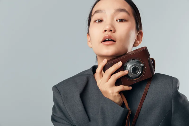 Young woman holding vintage camera close to face wearing oversized dark jacket posing against plain background. Female model with retro photo equipment and casual style for portrait photography
