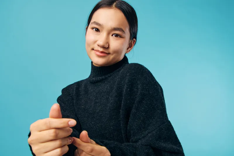Young woman with black hair wearing dark sweater smiling confidently against blue background with hand gesture in closeup studio portrait, beauty and casual fashion.