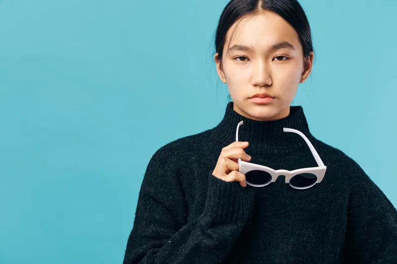 Young woman in black sweater holding white sunglasses looks at camera with neutral expression on blue background. Casual style, minimal fashion and portrait with natural light.