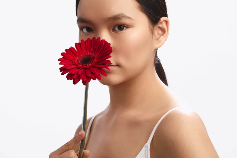 asian woman holds a vivid red flower near her face in a clean studio setting, soft lighting and gentle expression create a calm, intimate portrait with grace.