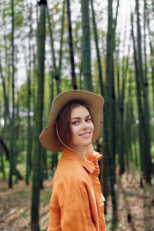 Woman portrait in bamboo forest, smile and wearing a straw hat with orange jacket, nature outdoor scene, relaxed pose and natural sunlight filtering through tall trunks.