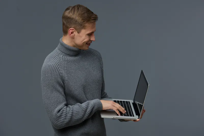 young smiling caucasian man wearing grey turtleneck sweater hold laptop look screen typing on keyboard isolated on dark grey background digital nomad concept