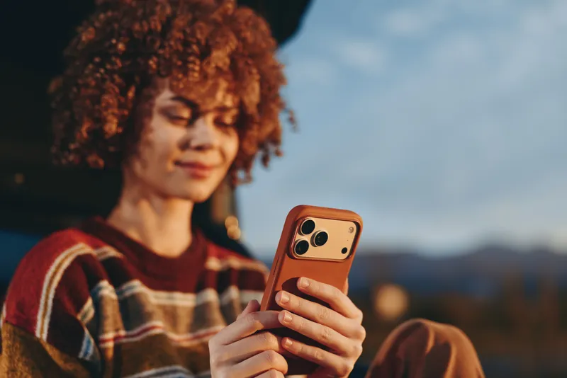 Woman with curly hair outdoors holding a smartphone in her hands, soft portrait framing against blue sky, lifestyle moment of communication, casual everyday routine, warm natural light