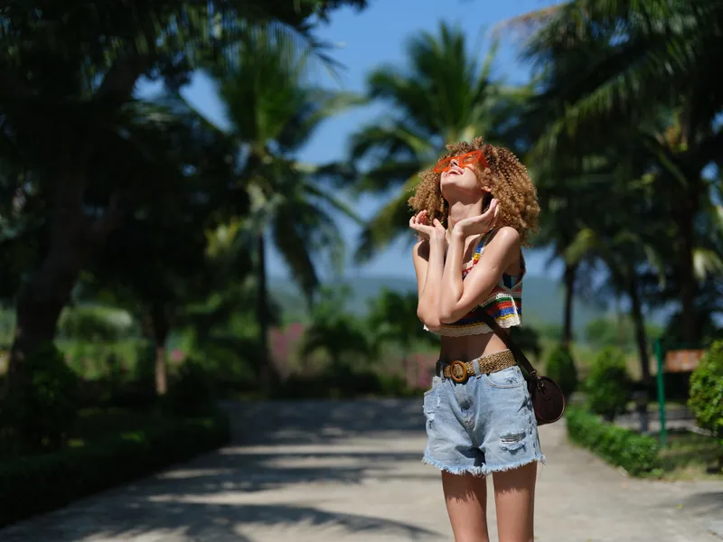 Young woman enjoying summer outdoors in casual denim shorts and colorful top, surrounded by tropical palm trees and bright sunlight.