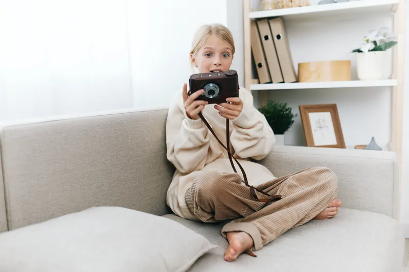 A young child sits cross legged on a soft sofa, holding a vintage camera and exploring photography in a cozy home setting with warm light and calm ambience.