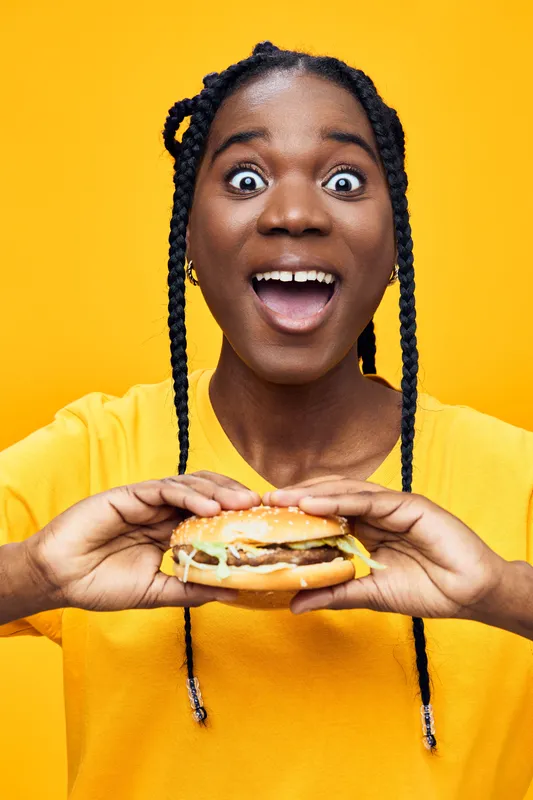 Excited young woman with braids holding a delicious burger against a bright yellow background, showcasing joy and enthusiasm for fast food