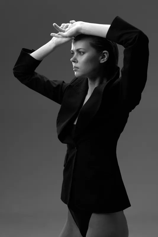 Black and white portrait of a thoughtful woman in a dark jacket with raised arms posing against a plain background.