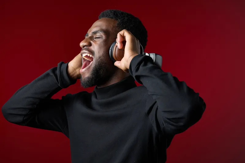 Young Black man joyfully singing with headphones against a bold red background, expressing happiness and passion for music