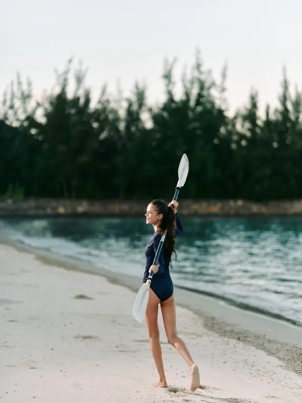 Young girl stands on a sandy beach in a swimsuit, lifting a paddle above her head while a second paddle rests by her side, ready for a sea adventure.