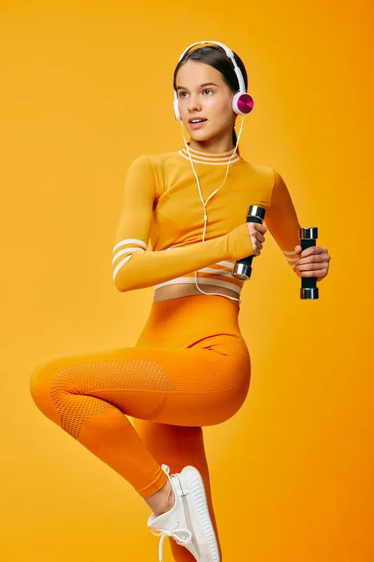 Woman fitness portrait in orange activewear, holding dumbbells with headphones, balancing on one leg against a bright studio backdrop, dynamic sport pose and energetic workout movement
