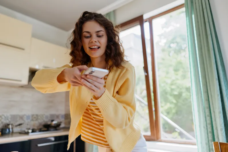 Young woman in a cozy kitchen using a smartphone, smiling, casual outfit, capturing a lifestyle moment at home, bright natural light, warm colors, relaxed daily routine.