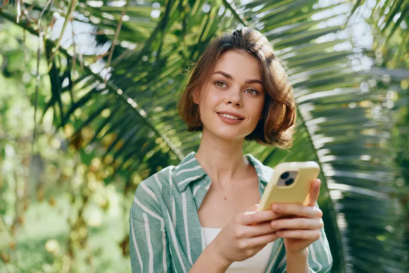 Young woman outdoors texting on a smartphone, smiling softly among vibrant tropical greenery.