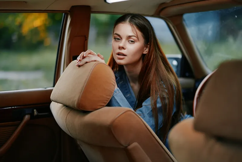 Young woman in a vintage car, showcasing natural beauty and confidence, dressed in casual denim, surrounded by a soft, warm background of autumn foliage.