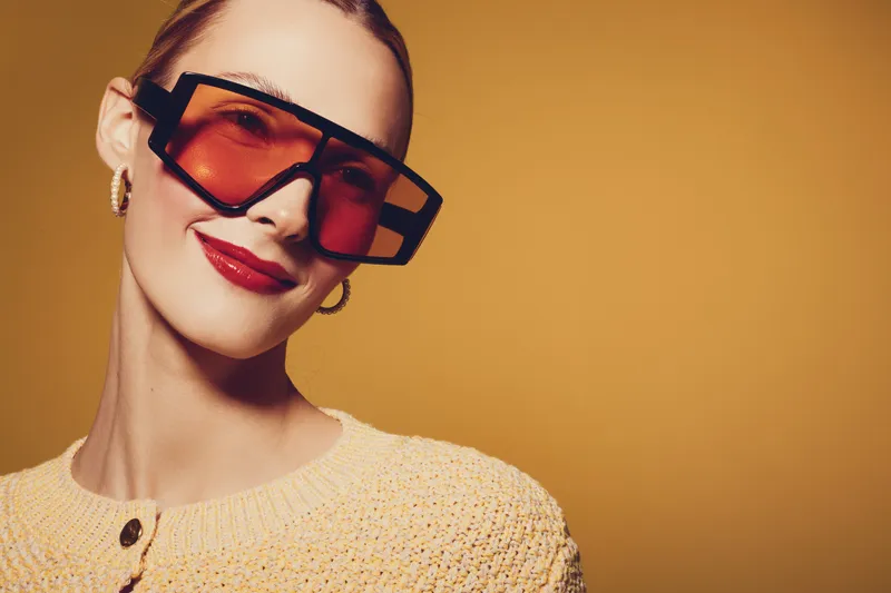 Smiling woman with red lipstick wearing oversized orange sunglasses and yellow knitted sweater. Studio portrait on warm brown background. Concept of fashion, style, and confident beauty.