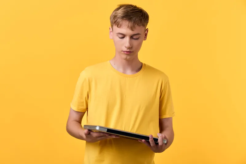 Young man in a yellow t shirt holds a tablet, concentrating on the screen in a bright studio, illustrating digital learning, online reading, and casual tech use.