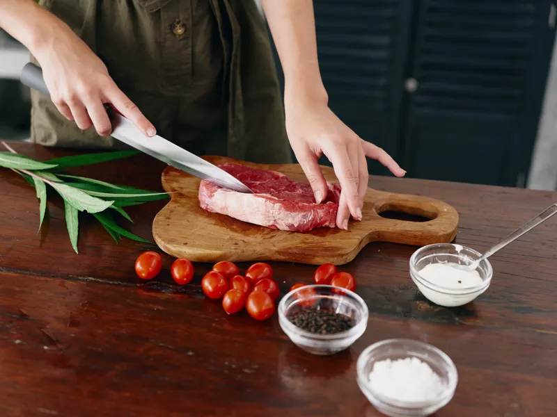 Женщина с ножом в руке нарезает свежий стейк для запекания на кухне, на столе соль, перец и другие специи, красные помидоры черри и зелень, приготовление ужина