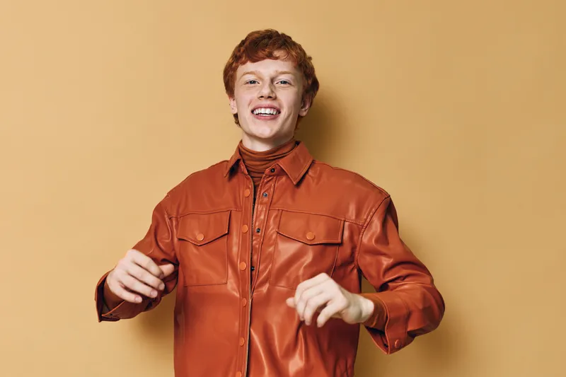 Young man with red hair wearing orange leather shirt laughs joyfully with open mouth and raised hands on beige background. Casual style, happiness and carefree mood in studio portrait.